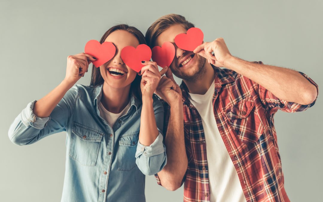 A young man and a woman hold paper cutouts of hearts over their eyes and smile.