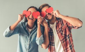 A young man and a woman hold paper cutouts of hearts over their eyes and smile.